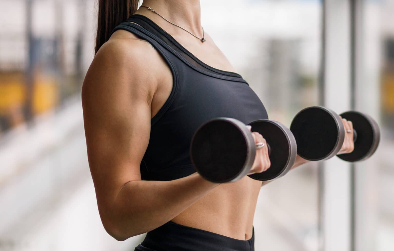 Young athletic fitness girl doing biceps curls with dumbbells near the window